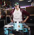 A person wearing sunglasses and a white subway sleeve t-shirt with a graphic design, sitting in front of a drum set in a store.