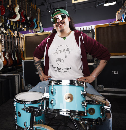 A person wearing sunglasses and a white subway sleeve t-shirt with a graphic design, sitting in front of a drum set in a store.