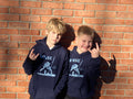 Two children wearing navy blue hoodies with a logo against a brick wall.