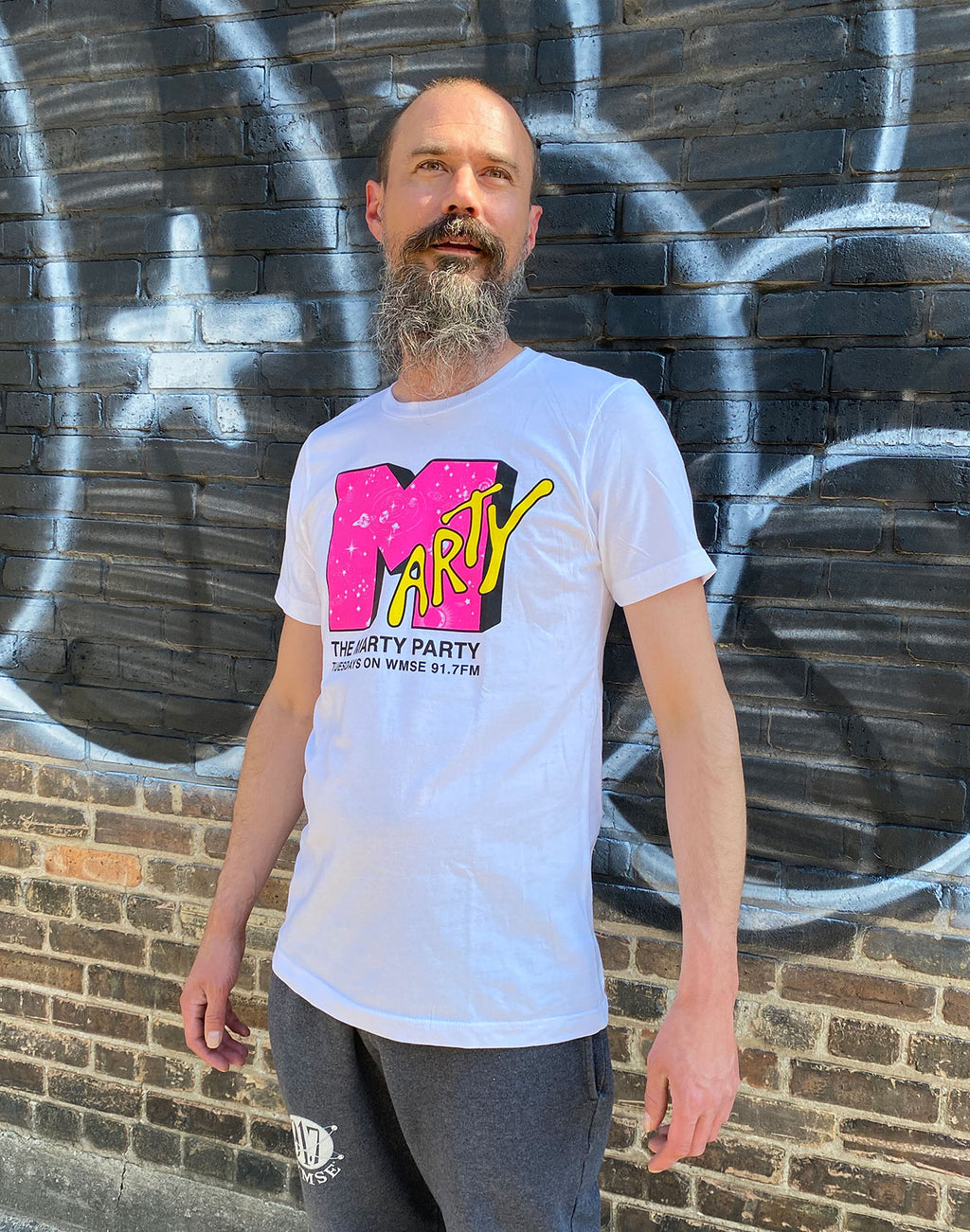 Man wearing a white t-shirt with colorful graphics against a graffiti-covered wall.