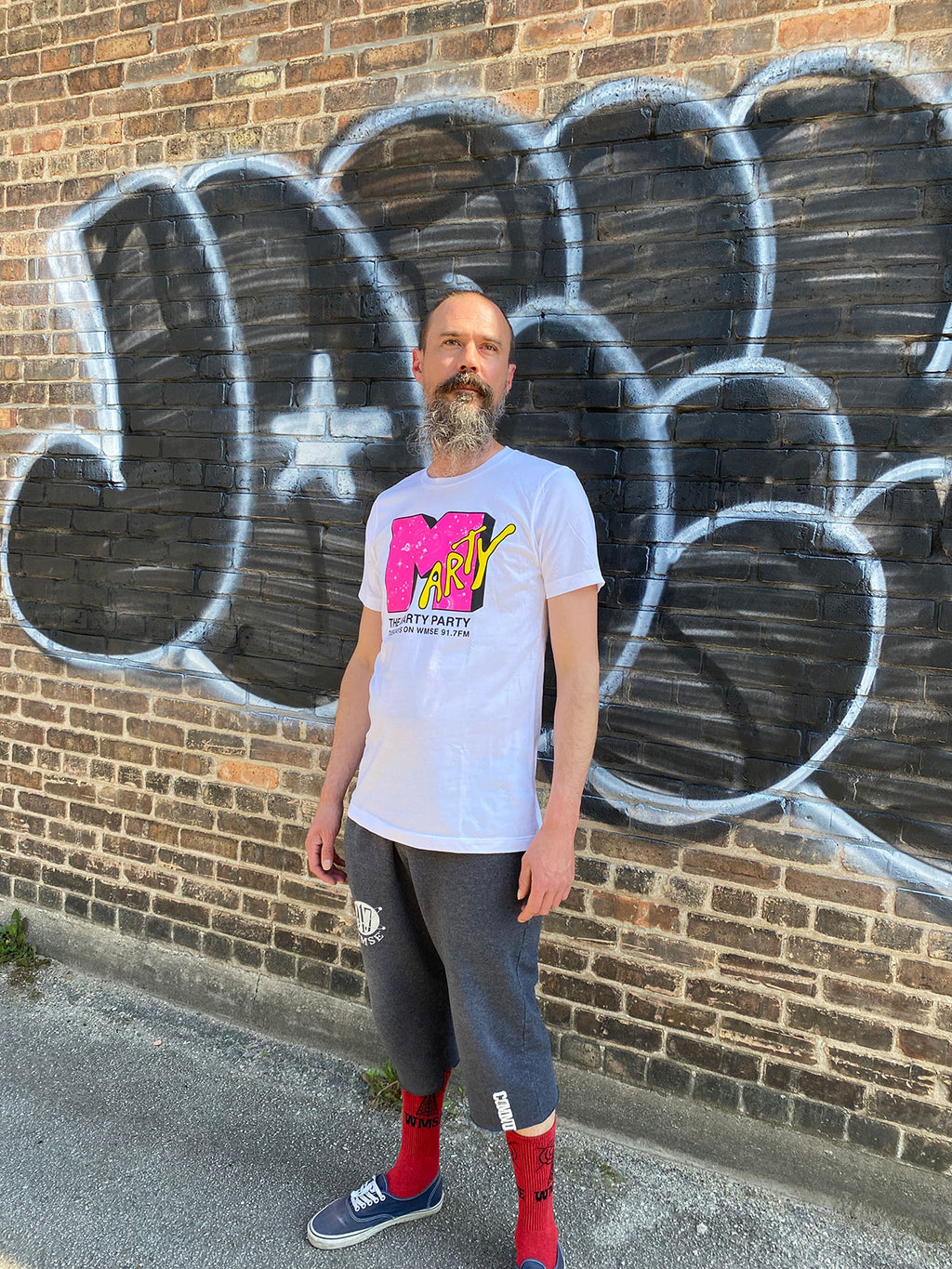 Man wearing a white t-shirt with colorful design standing in front of a brick wall with graffiti.